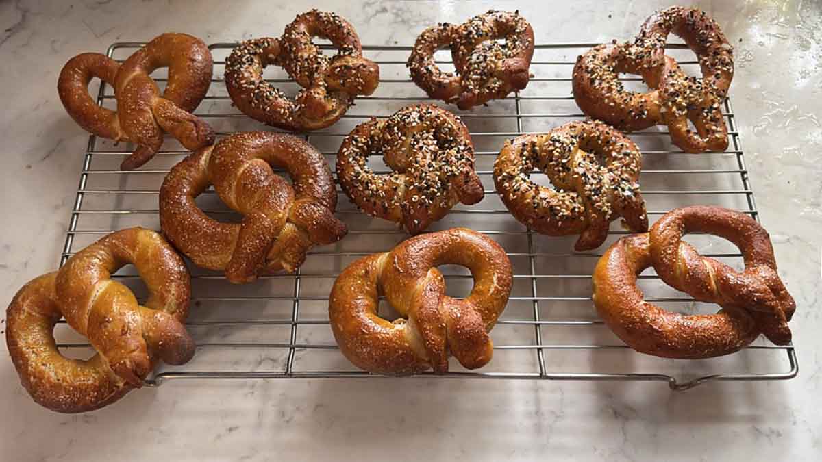 Ten homemade soft pretzels, half plain the other half coated with white and black sesame seeds cooling on a wire rack.