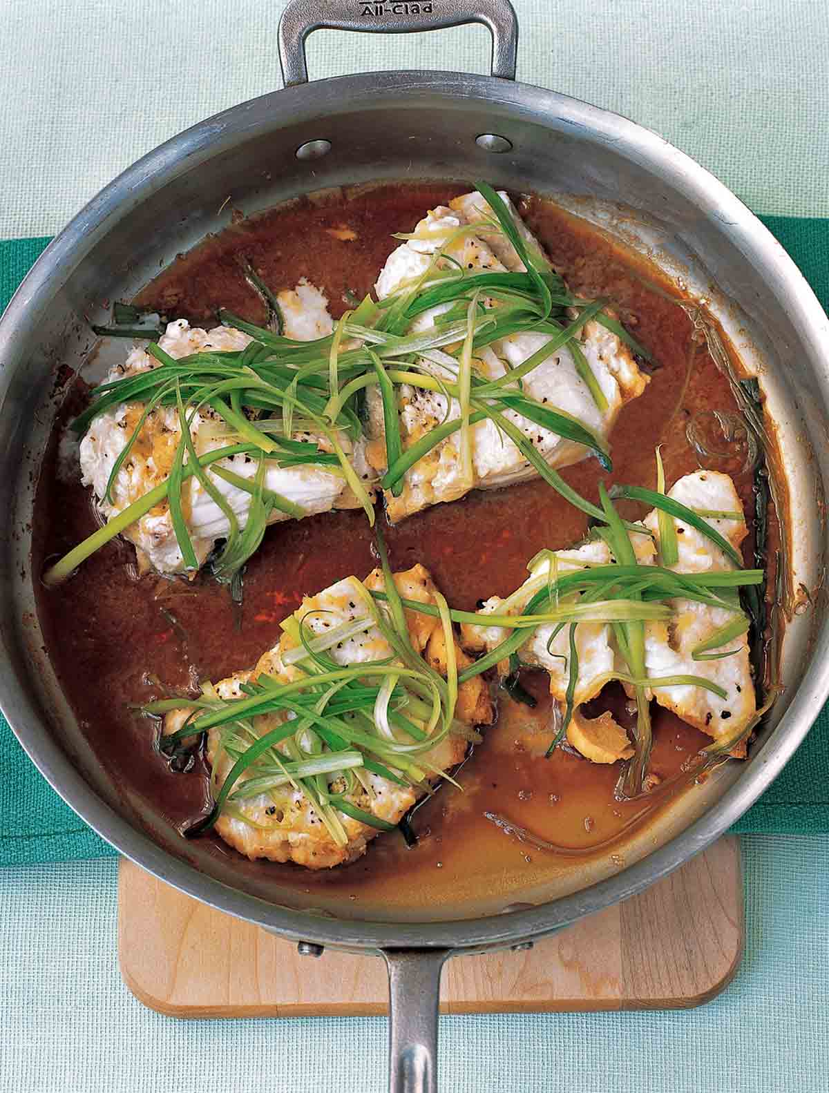 A metal skillet with four pieces of steamed cod with ginger and scallions.