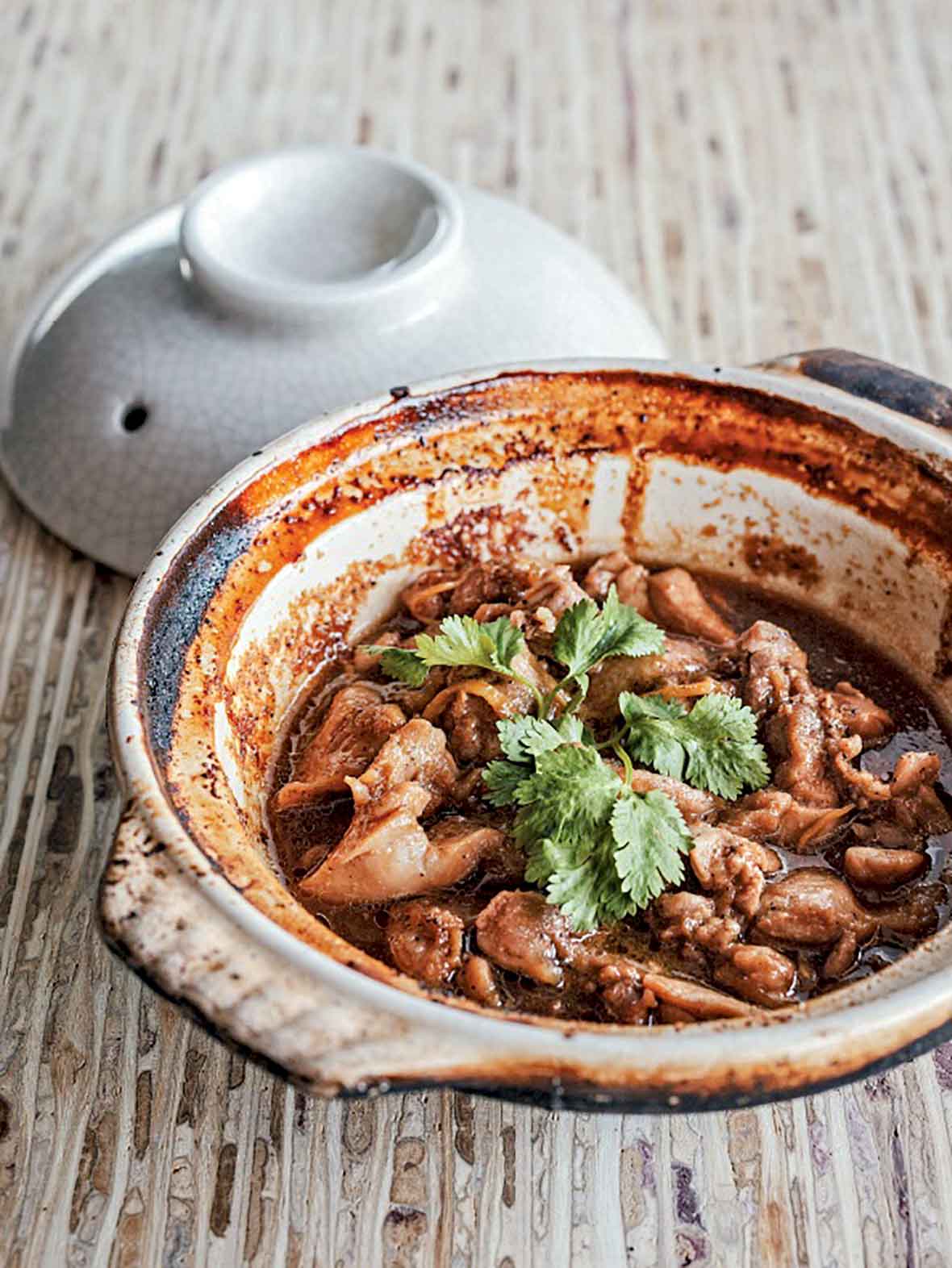 Clay pot filled with Vietnamese caramel chicken pieces and cilantro on a wooden table.