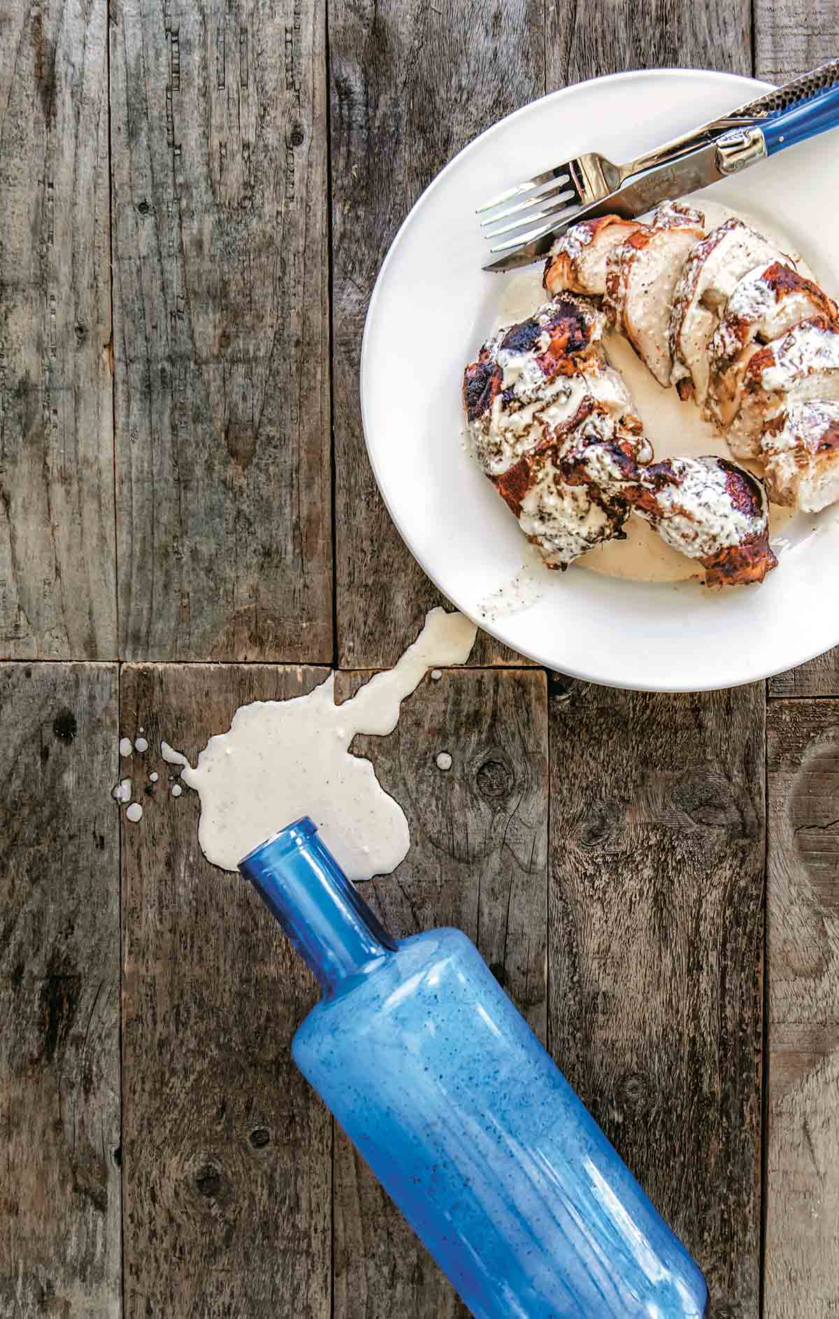 A blue bottle of Alabama white bbq sauce spilled on a wooden table with a plate of grilled chicken covered in white bbq sauce beside it.
