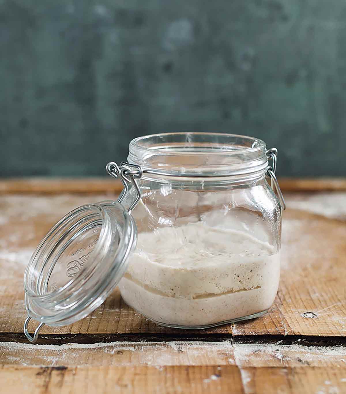 A glass jar with growing starter on a wooden surface as part of an explanation on how to make sourdough starter.