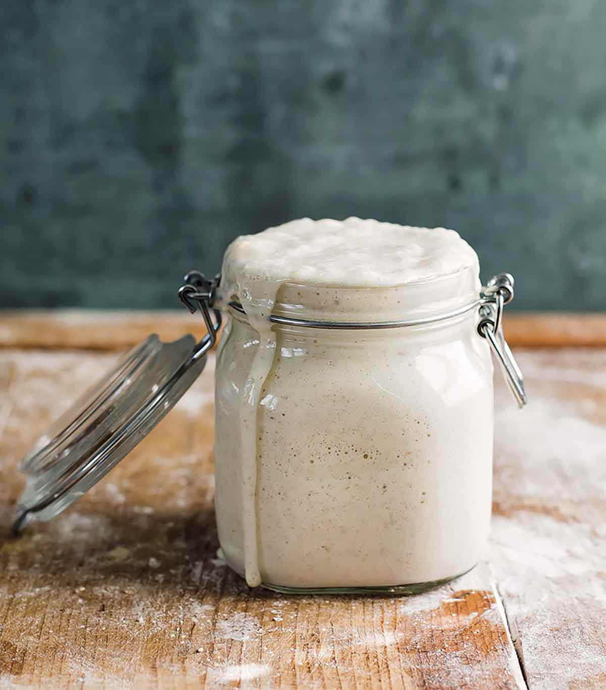 A glass jar with overflowing starter on a wooden surface as part of an explanation on how to make sourdough starter.
