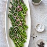 A white oval platter filled with asparagus with raspberry-shallot vinaigrette, and pickled shallots scattered on top.