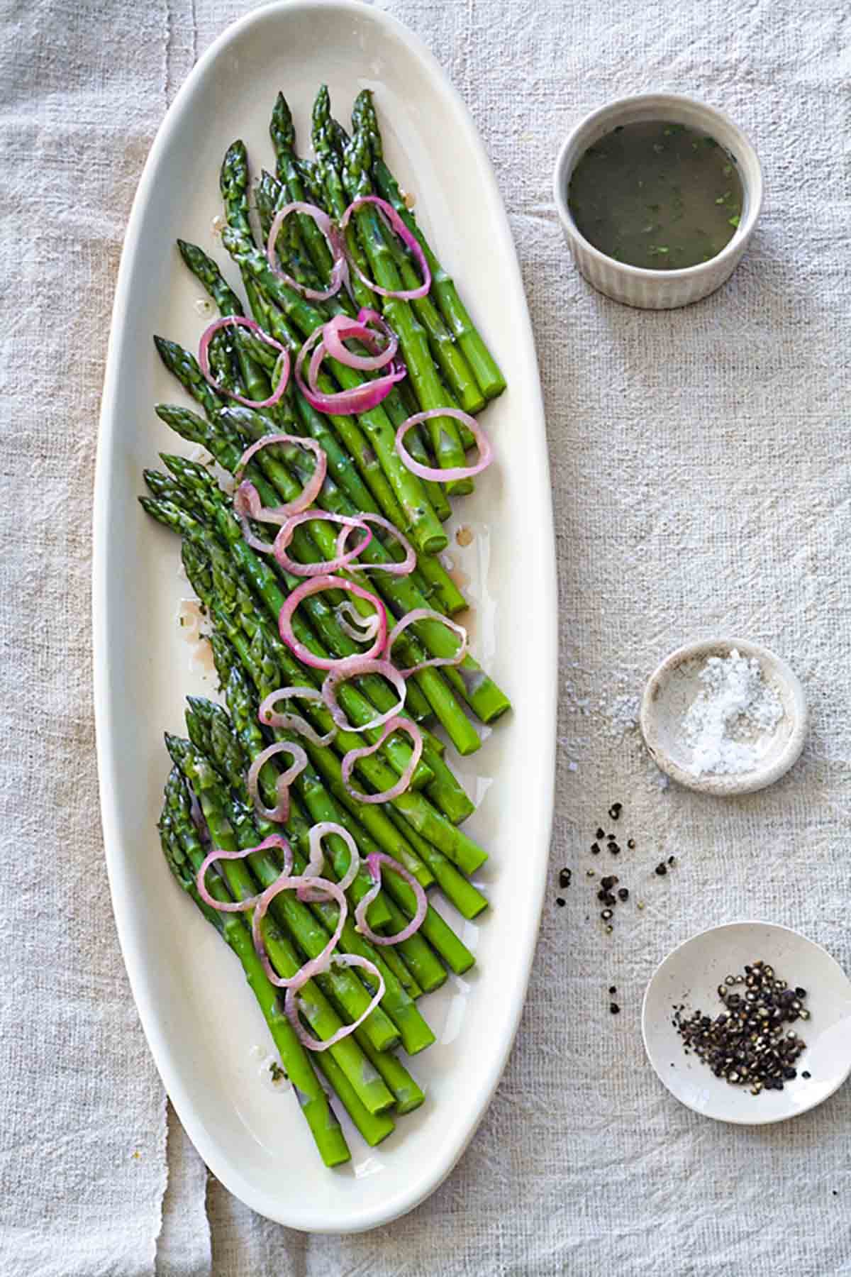 A white oval platter filled with asparagus topped with a raspberry and shallot vinaigrette and pickled shallots.