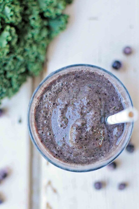 An overhead view of a glass of blueberry kale smoothie with a straw and some blueberries and kale scattered around the glass.
