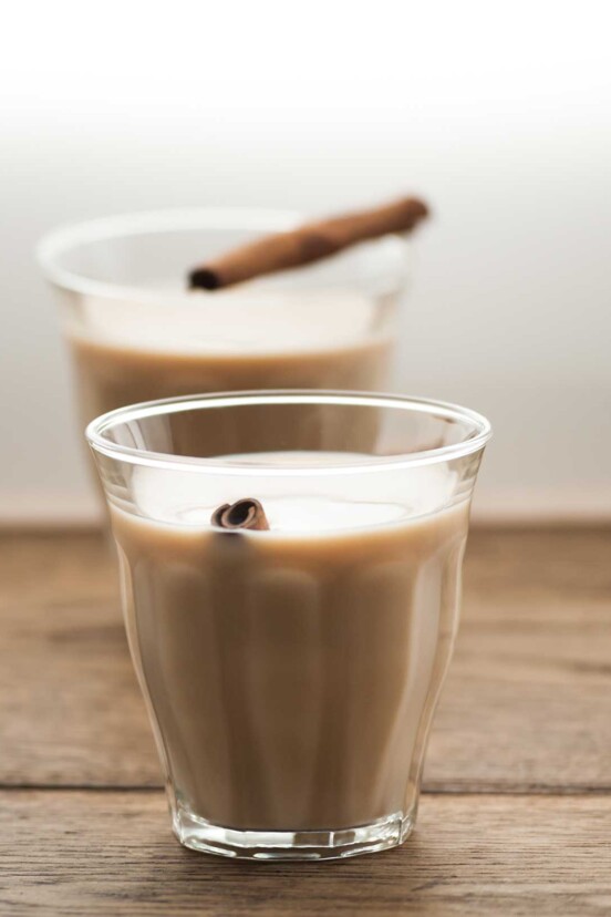 Two glasses of chai latte with cinnamon sticks on a wooden surface.