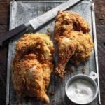 Two pieces of cornmeal-crusted fried chicken with white barbecue sauce and a knife on a metal sheet.