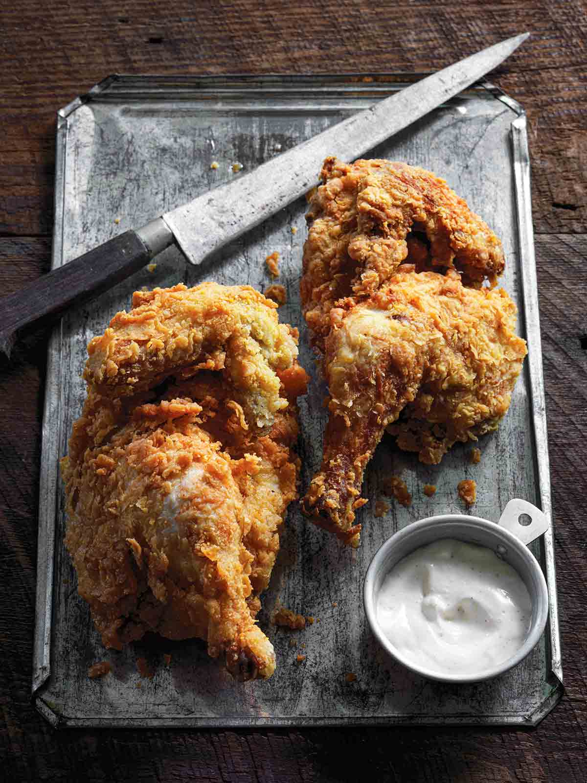 Two pieces of cornmeal-crusted fried chicken with white barbecue sauce and a knife on a metal sheet.