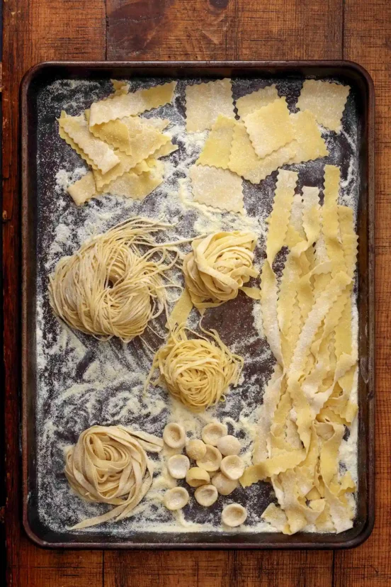 A sheet pan of homemade egg pasta in different shapes: ruffled mafaldine ribbons, rectangular sagnarelli, nests of tagliatelle and spaghetti, and ear-shaped orecchiette.