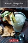 A tall glass of frozen margarita with a salt rim and lime wheel beside a bowl of chips and guacamole