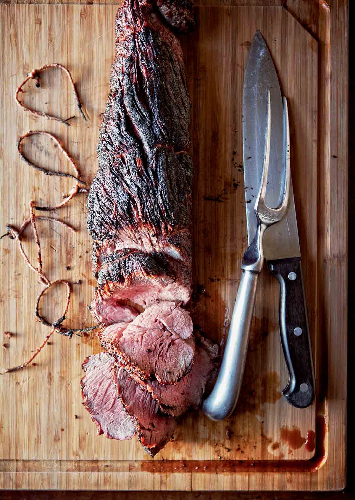 A whole grilled beef tenderloin, partially sliced on a wooden cutting board with a knife, meat fork, and twine beside it.