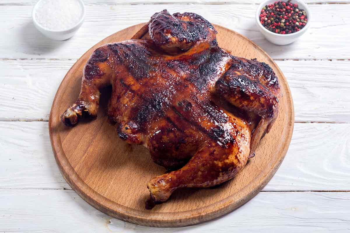 A whole spatchcocked grilled roasted chicken on a round wooden board with dishes of salt and peppercorns on the side.
