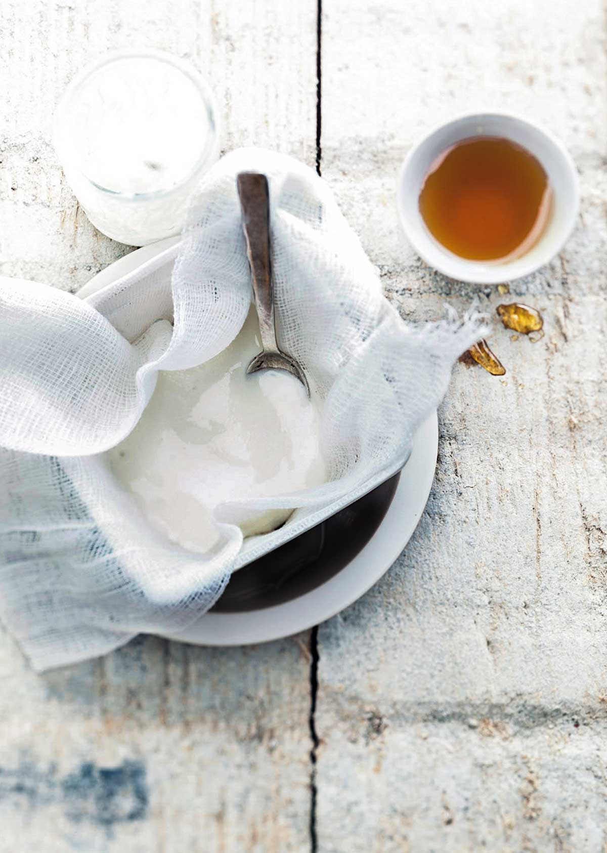 Homemade Greek yogurt being strained through cheesecloth into a white bowl with a spoon and a small bowl of honey beside it.