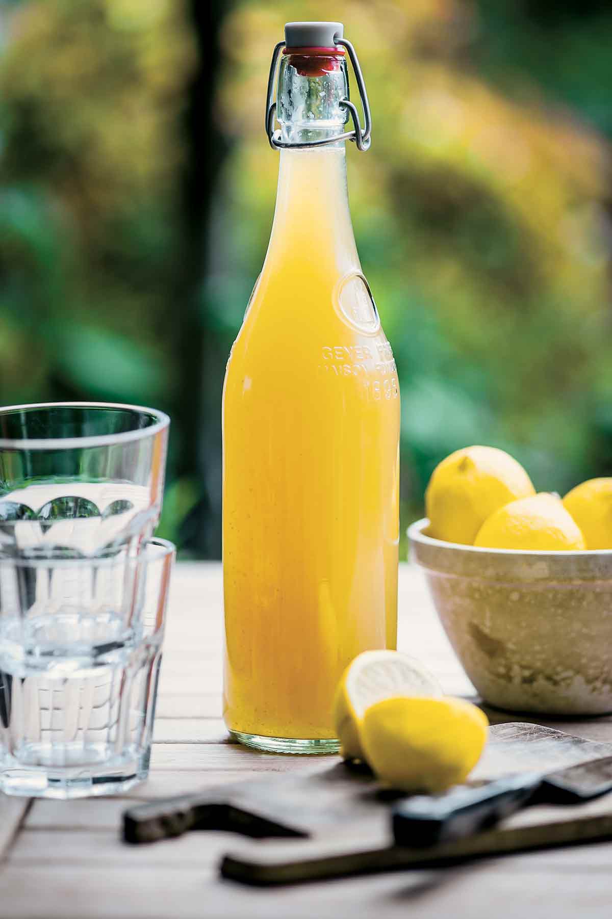 En glasflaska fylld med hemgjord lemonadsirap, en skål med citroner och två glas på ett träbord.