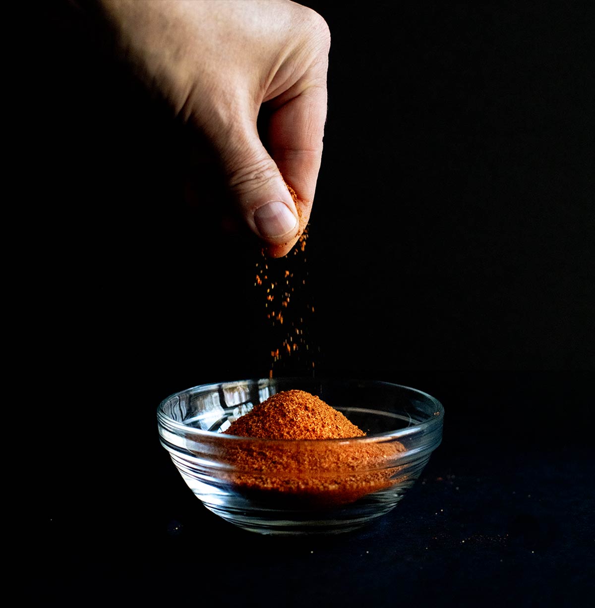 A hand sprinkling homemade Old Bay Seasoning in a small glass bowl