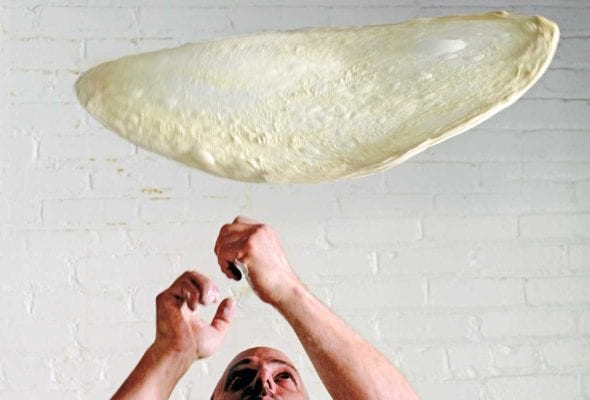 A thin circle of stretched pizza dough being tossed in the air.