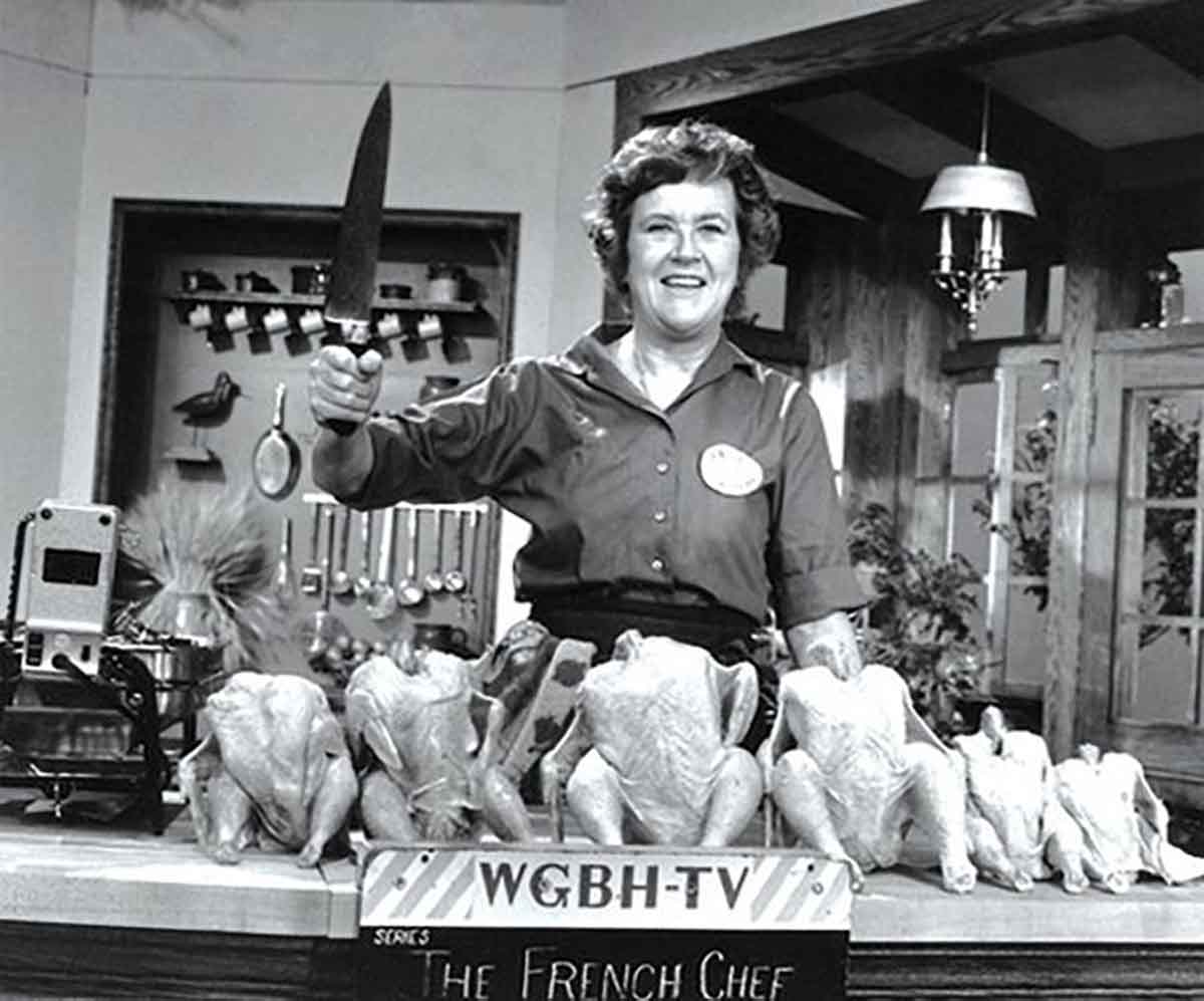 Julia Child on set with knife in the air and chickens on the counter.