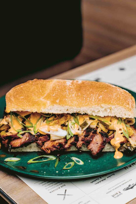 A kimchi bulgogi sandwich with sliced steak, green onions, and mayo on a roll on a green plate on top of a menu.
