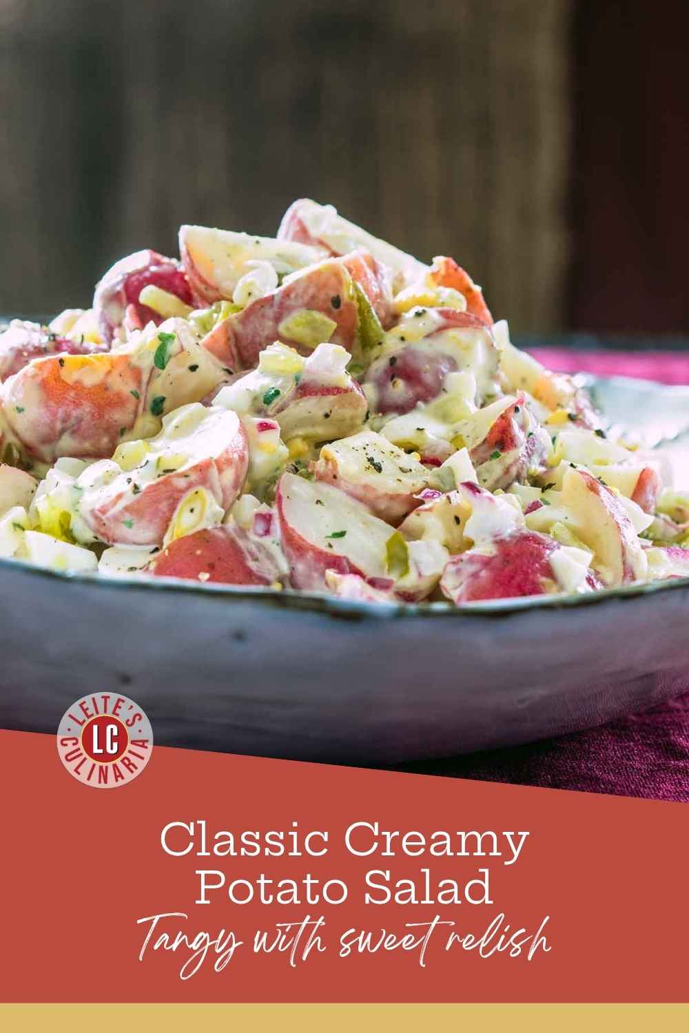 Chunky red potato salad with hard-boiled eggs and sweet relish in a grey ceramic bowl on a burgundy tablecloth.
