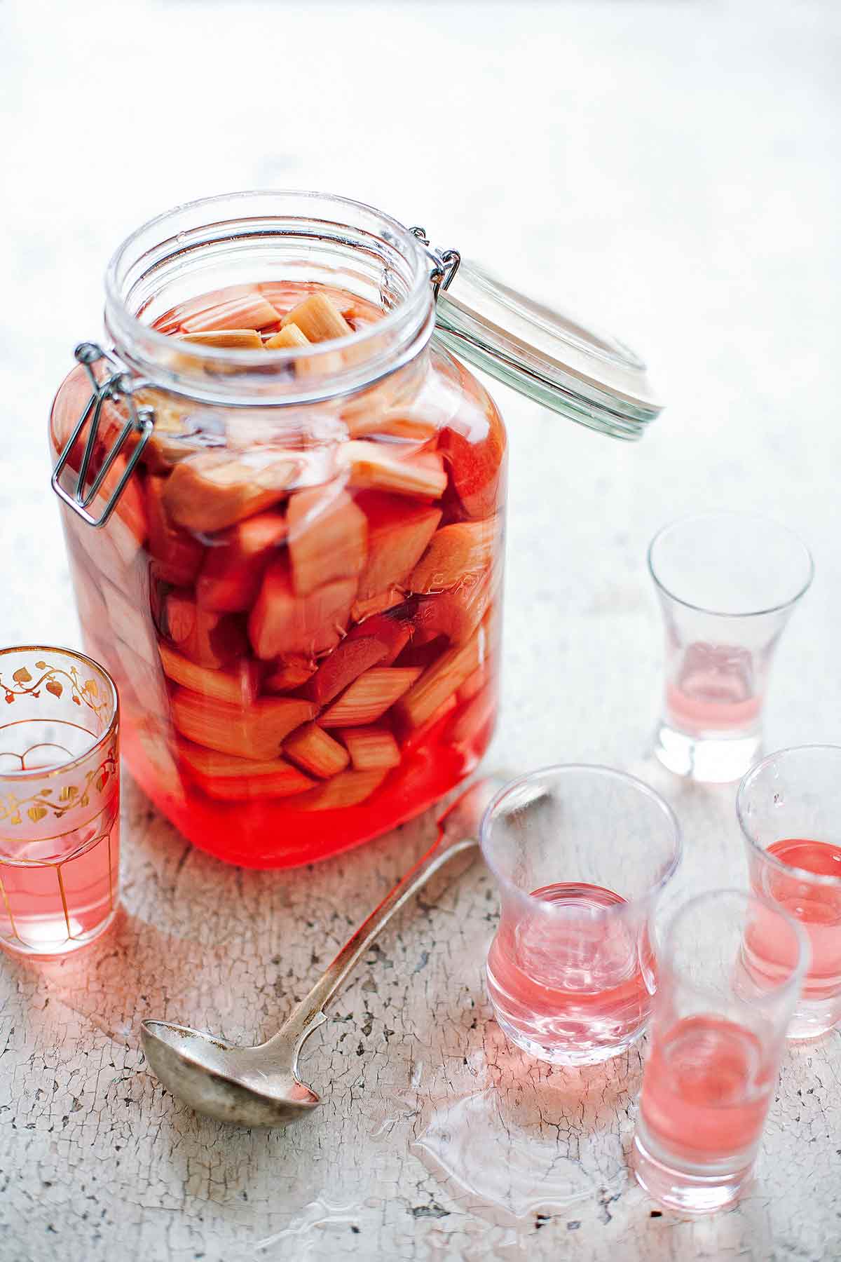 A canning jar filled with rhubarb vodka and pieces of rhubarb with several crystal glasses partially filled with rhubarb vodka beside it.