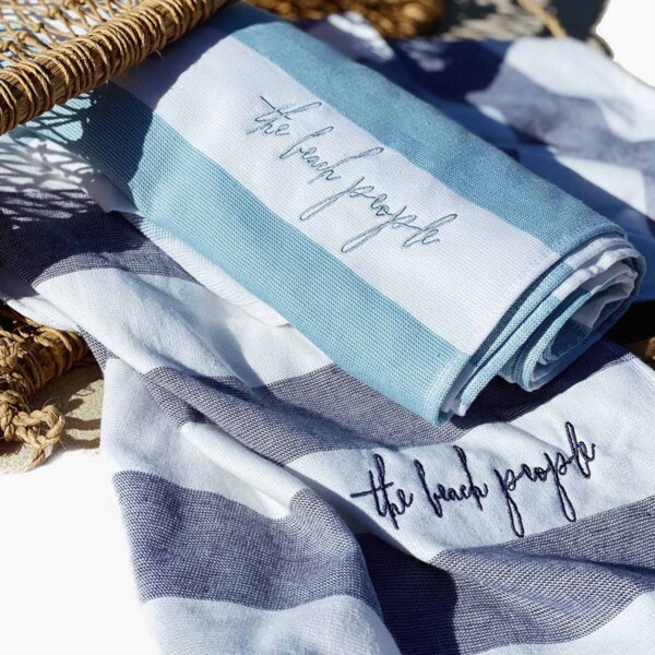 Two sand-free cabana towels inscribed with 'the beach people' lying beside a wicker basket.