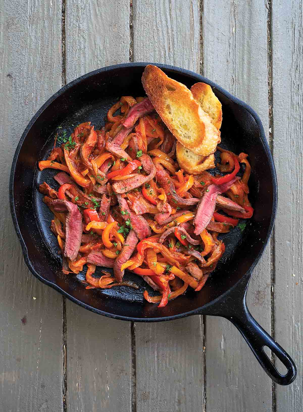 Skillet steak peperonata and two slices of toasted baguette in a cast-iron pan.