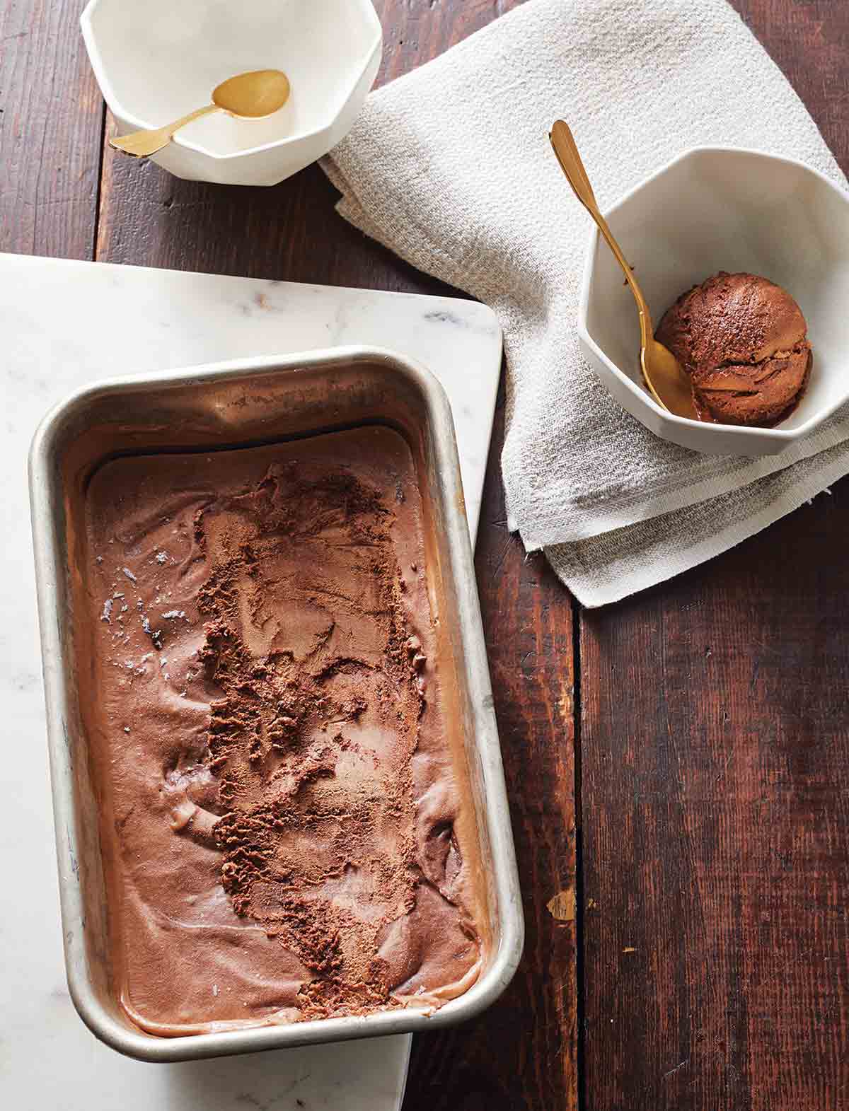 A metal dish filled with vegan chocolate fudge swirl ice cream, as well as two bowls with gold spoons, one empty and one with a scoop of ice cream.