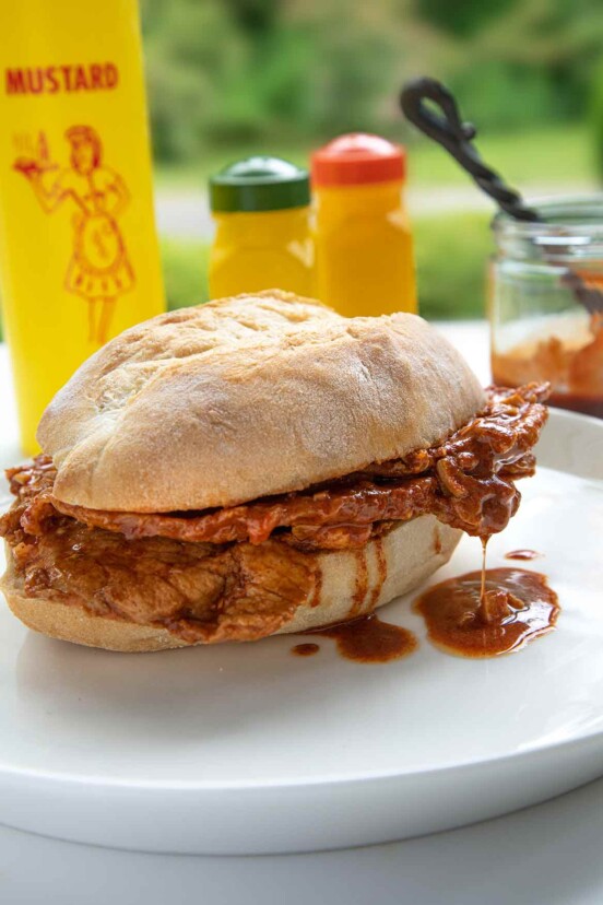 A bifana, a Portuguese pork sandwich on a white plate with a bottle of mustard in the background.