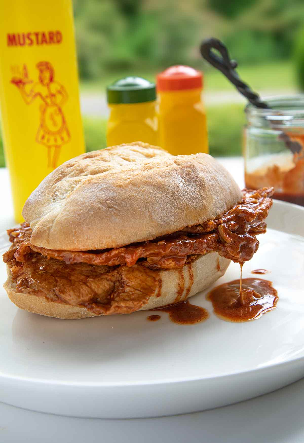 A bifana, a Portuguese pork sandwich on a white plate with a bottle of mustard in the background.