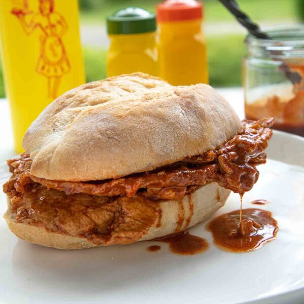 A bifana, a Portuguese pork sandwich on a white plate with a bottle of mustard in the background.