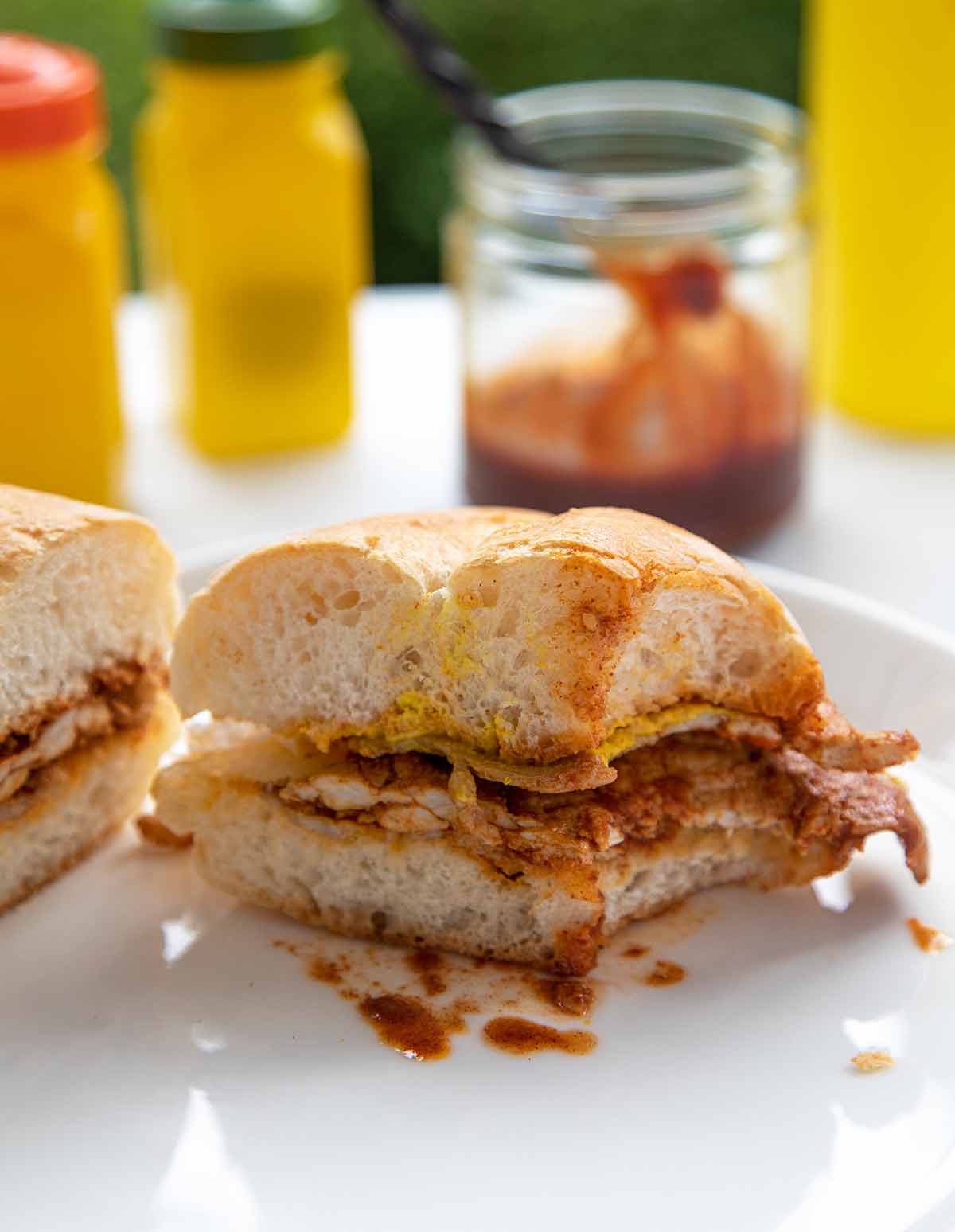 A bifana cut in half with a bite missing from it and a jar of piri-piri sauce in the background.