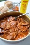 Thin slices of cooked pork in a pot for bifanas, with mustard, piri-piri sauce, and papo-secos in the background.