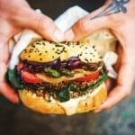 A person holding a black bean burger filled with spinach, tomato, onion, and asparagus.