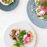 Three plates filled with shredded beef tacos--braised beef brisket, Cotija cheese, napa cabbage, sour cream, red onions, and cilantro on charred corn tortillas