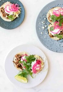 Three plates filled with shredded beef tacos--braised beef brisket, Cotija cheese, napa cabbage, sour cream, red onions, and cilantro on charred corn tortillas