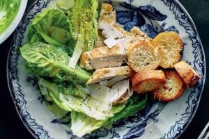 A blue and white plate topped with a deconstructed chicken Caesar salad with baguette round croutons.