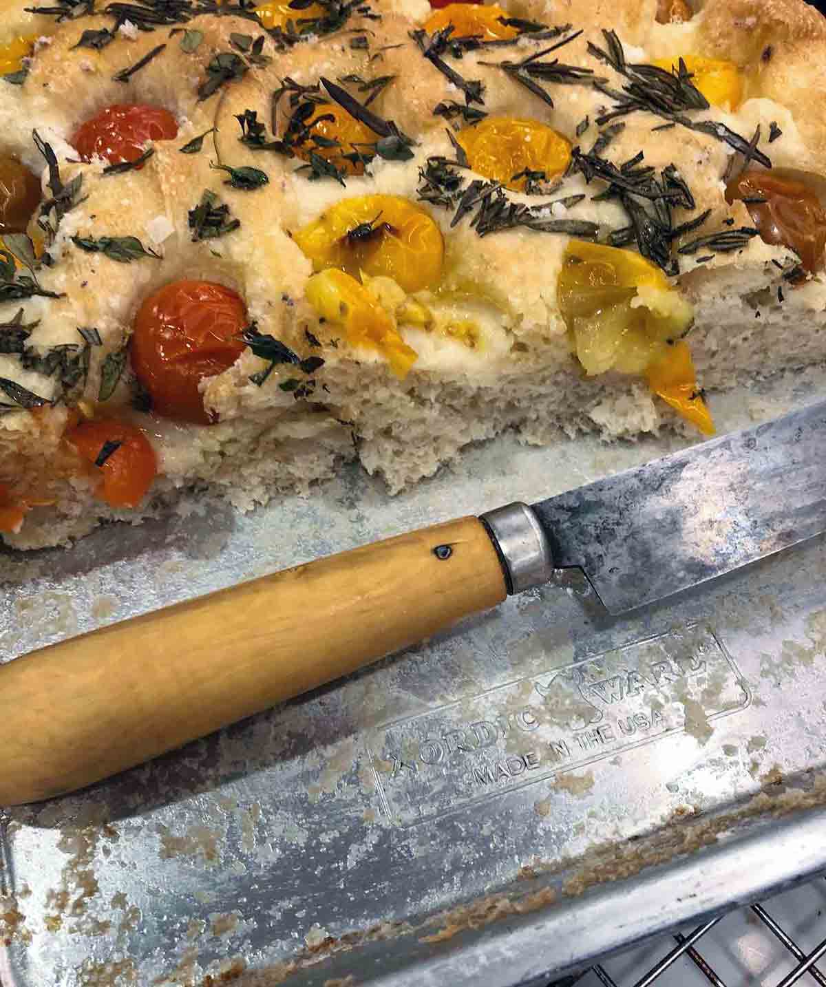 A cut loaf of red onion and rosemary focaccia with grape tomatoes on a rimmed baking sheet.