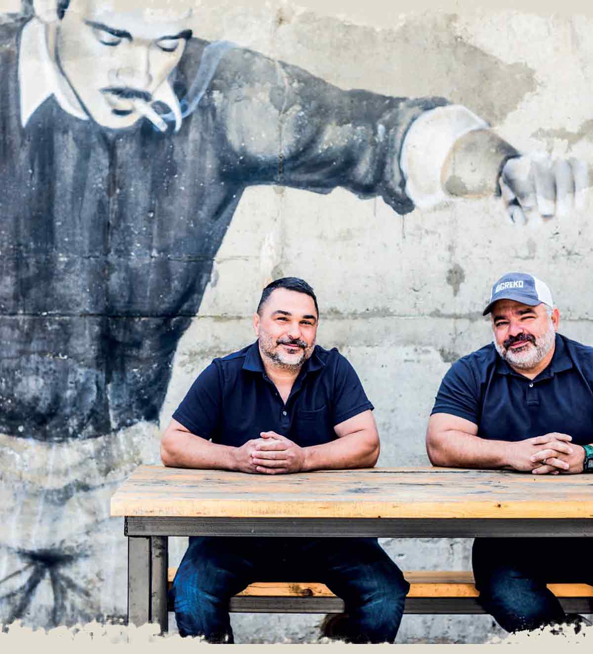 Greko chefs sitting at a wooden table in front of a wall mural.