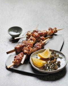 Four grilled pork skewers on a white plate with a bowl of lemon wedges and dried oregano beside the skewers.