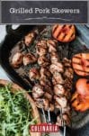 Four grilled pork skewers with peaches in a metal dish with a wooden bowl of arugula beside it.