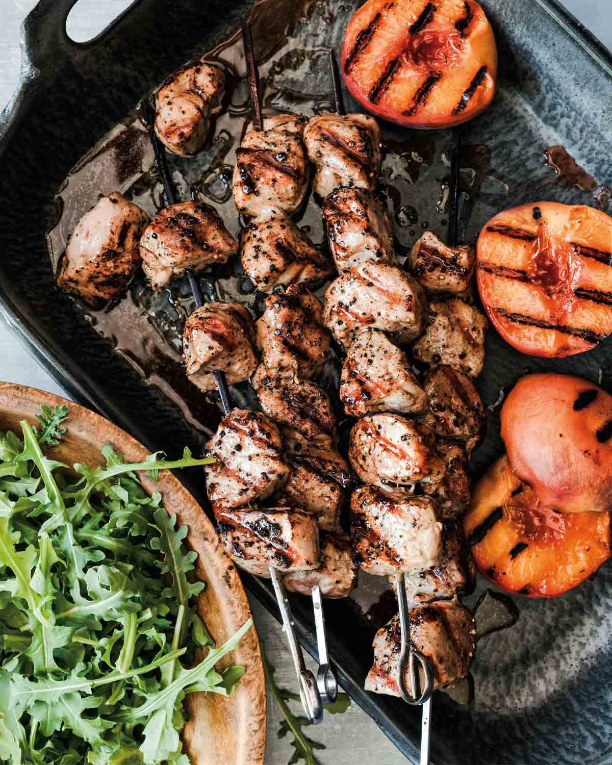 Four grilled pork skewers with peaches in a metal dish with a wooden bowl of arugula beside it.
