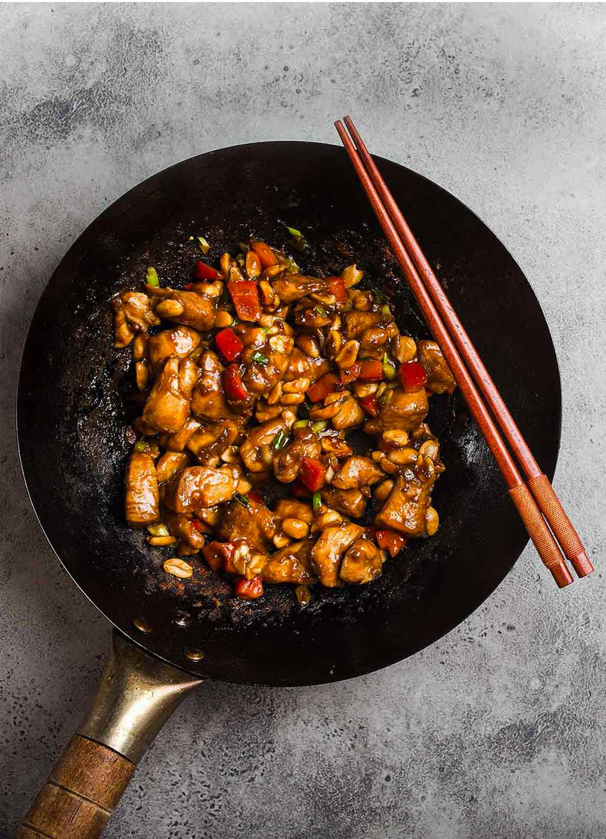 A wok filled with kung pao chicken with two chopsticks on the edge