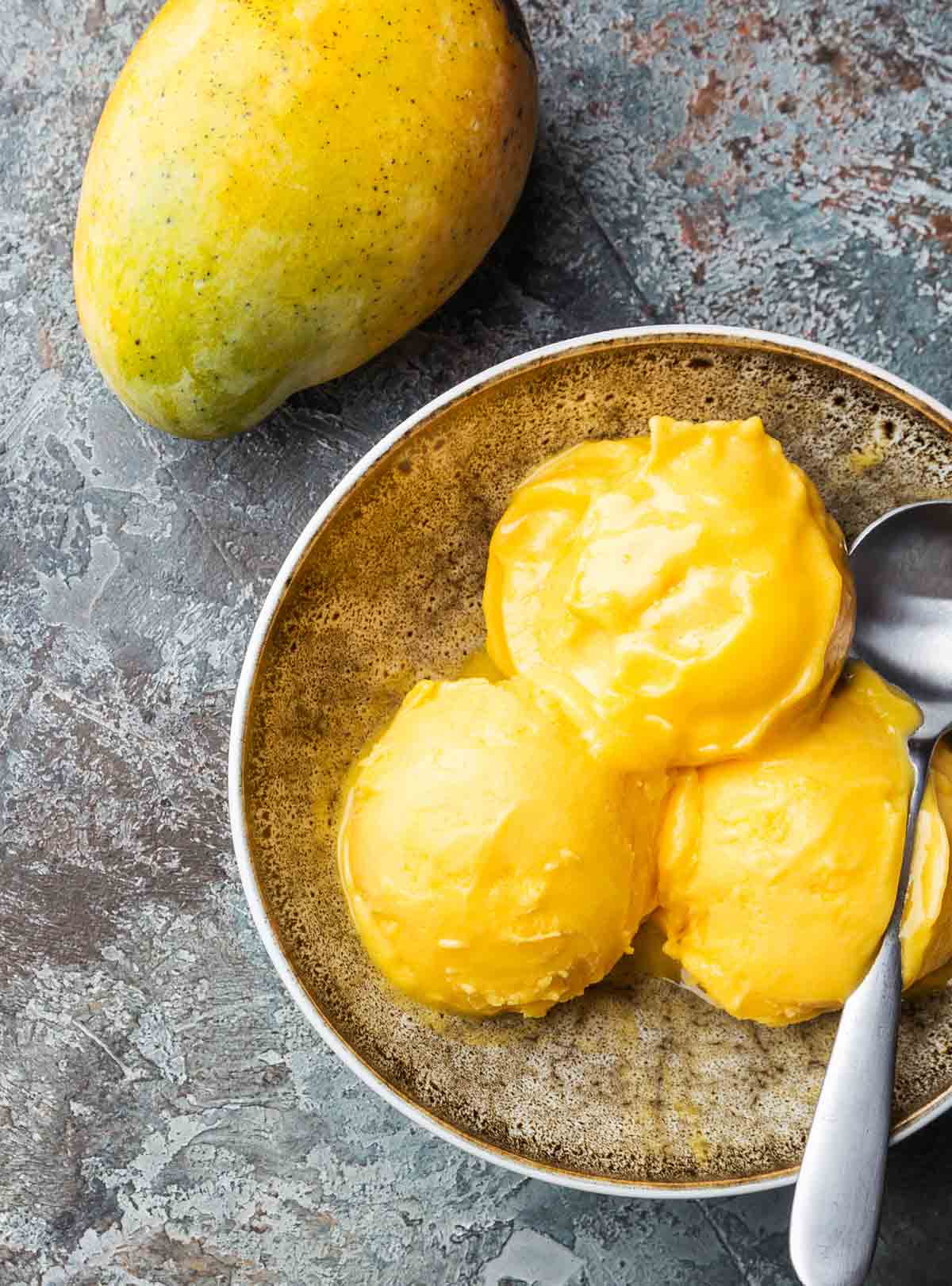 Three scoops of mango gelato in a bowl with a silver spoon and a whole mango beside it.