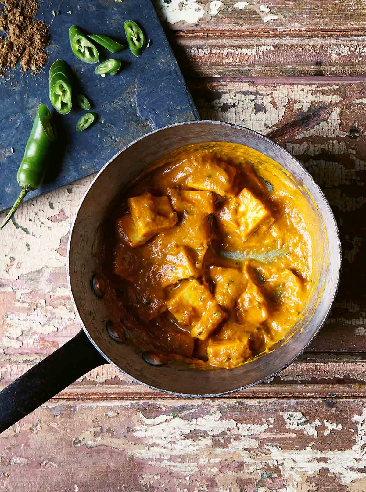 A saucepan filled with masala paneer on a wooden table with chopped jalapeno and spices on a blue board beside it.