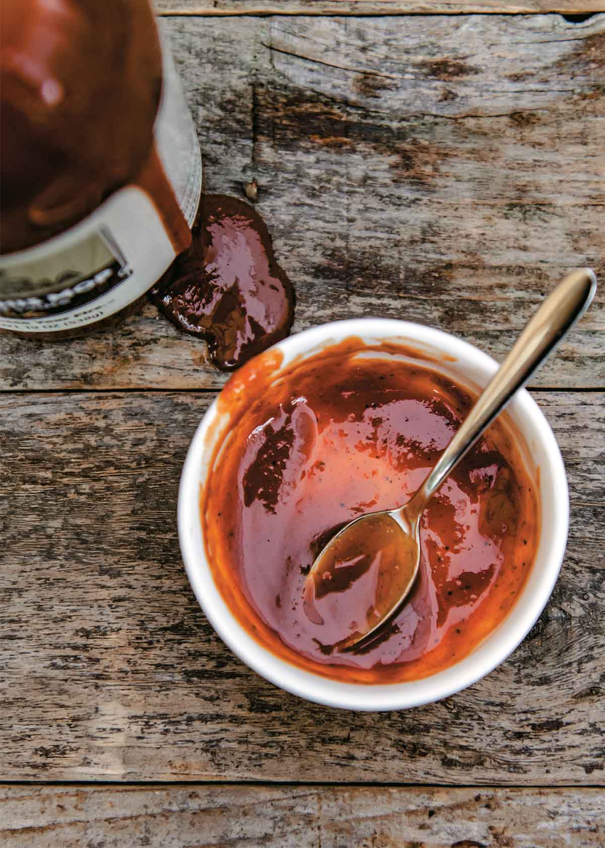 A white bowl with some Memphis BBQ mop sauce and a spoon in it with a bottle of sauce on the side.