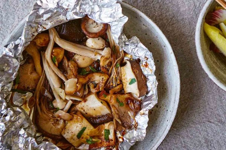 Two pouches of baked mushrooms in foil packets with a bowl of greens and another bowl of quinoa on the side.