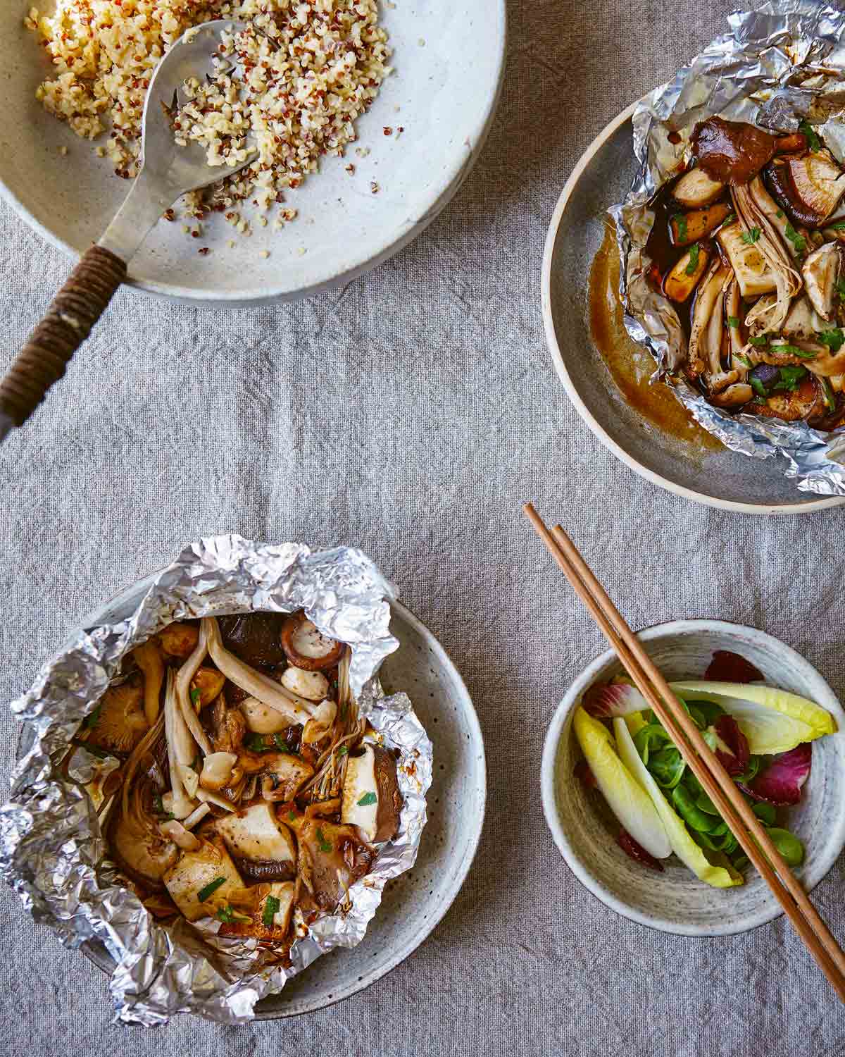 Two pouches of baked mushrooms in foil packets with a bowl of greens and another bowl of quinoa on the side.