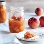 Two jars of peach and rhubarb jam with some peaches in the background and a plate with a piece of bread topped with the jam.