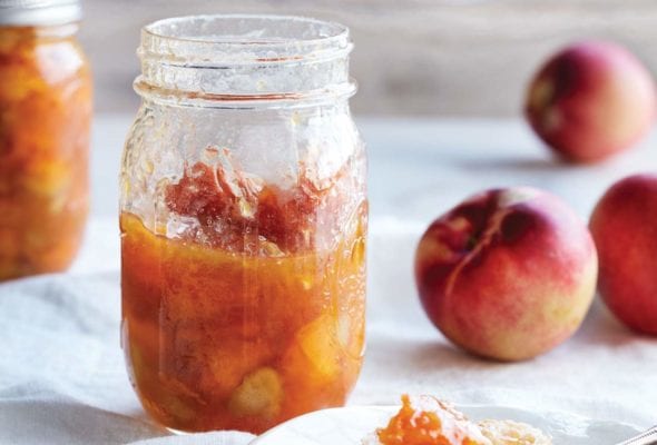 Two jars of peach and rhubarb jam with some peaches in the background and a plate with a piece of bread topped with the jam.