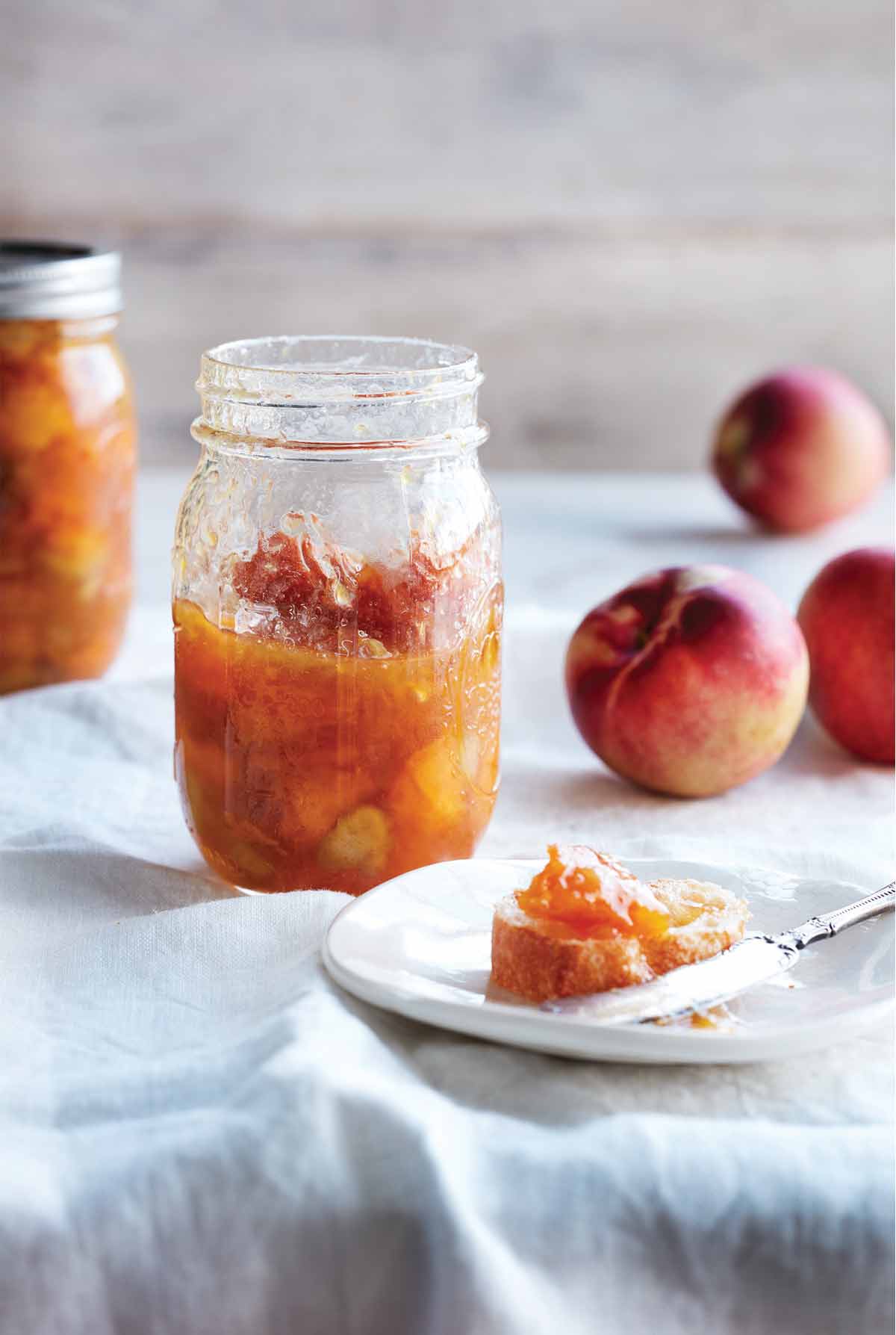 Two jars of peach and rhubarb jam with some peaches in the background and a plate with a piece of bread topped with the jam.